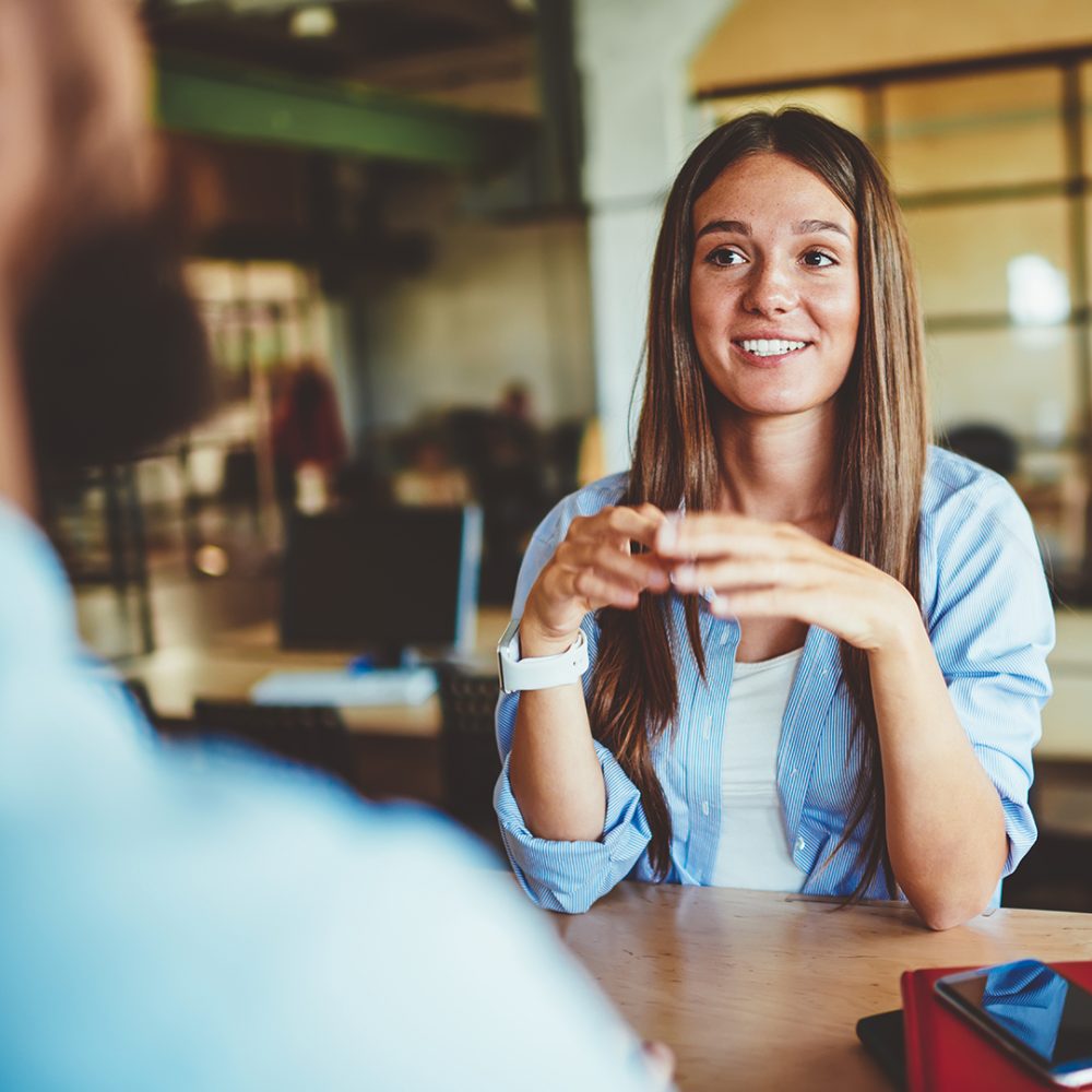 Positive smiling woman with brunette hair sitting at wooden table and talking with male friend in front, successful caucasian hipster girl casual dressed spending time with man and discussing ideas